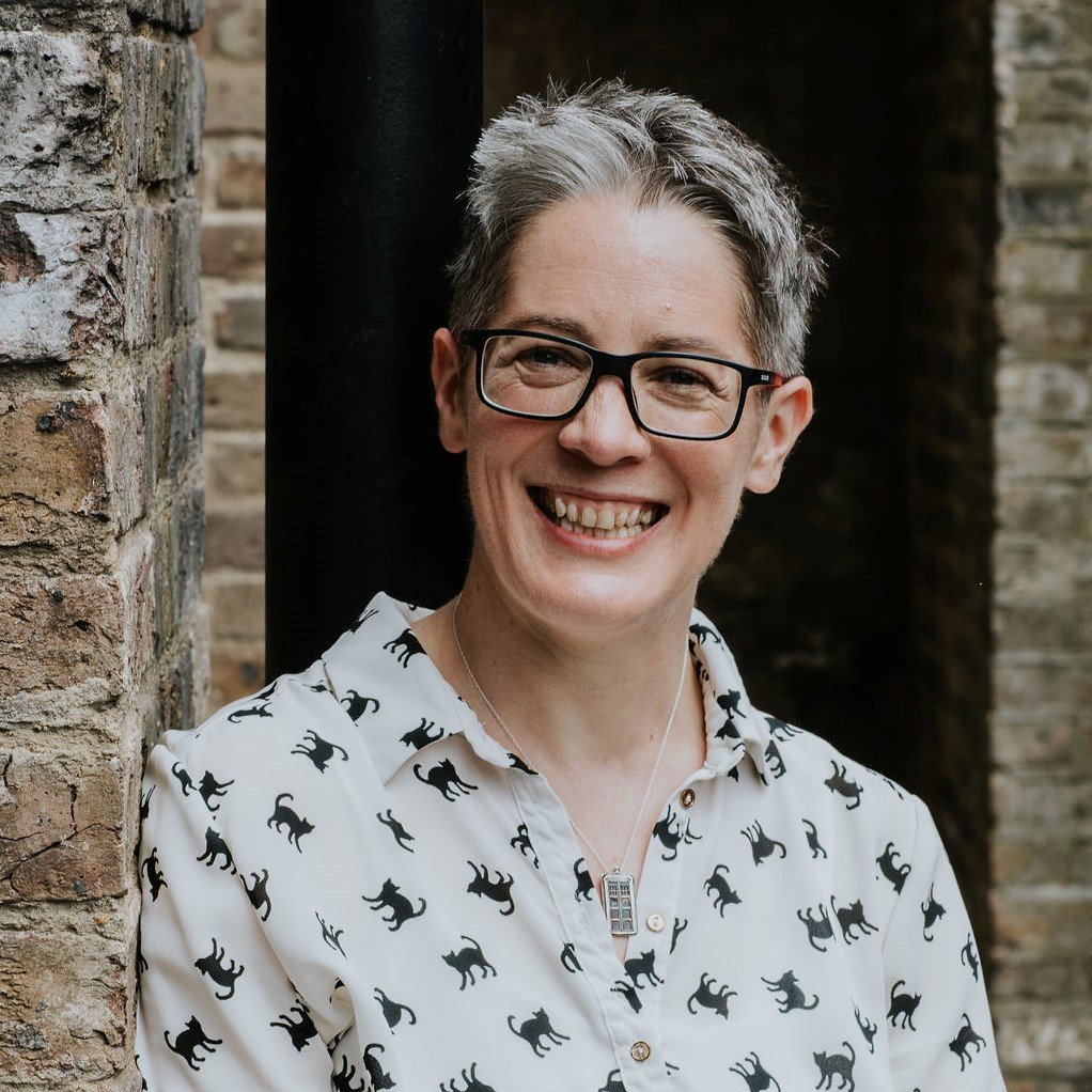 Jo Gibney, a White person with short hair and glasses, wearing a white shirt with black cats, leaning against a brick wall, smiling at the camera.